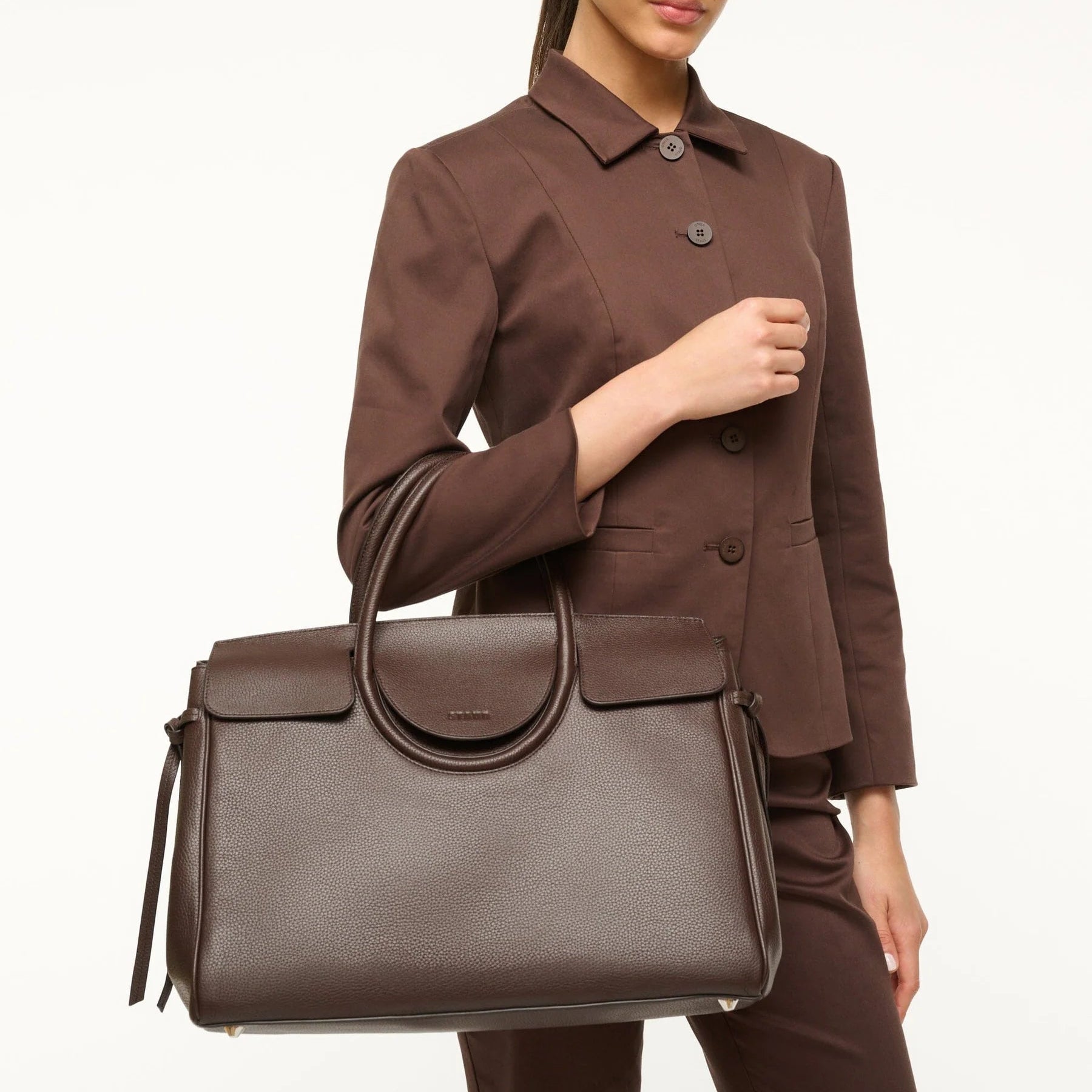 A woman in a brown suit holds the Staud Maude Carryall, an oversized bag crafted from structured dark brown pebbled leather, by its handles against a plain background.