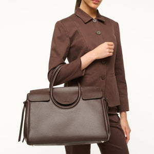 A woman in a brown suit holds the Staud Maude Carryall, an oversized bag crafted from structured dark brown pebbled leather, by its handles against a plain background.
