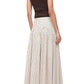 A person stands with their back to the camera, wearing a sleeveless dark brown top and the Staud Procida Skirt—a high-waisted white and beige striped maxi skirt with a curved yoke—paired with brown flat sandals.