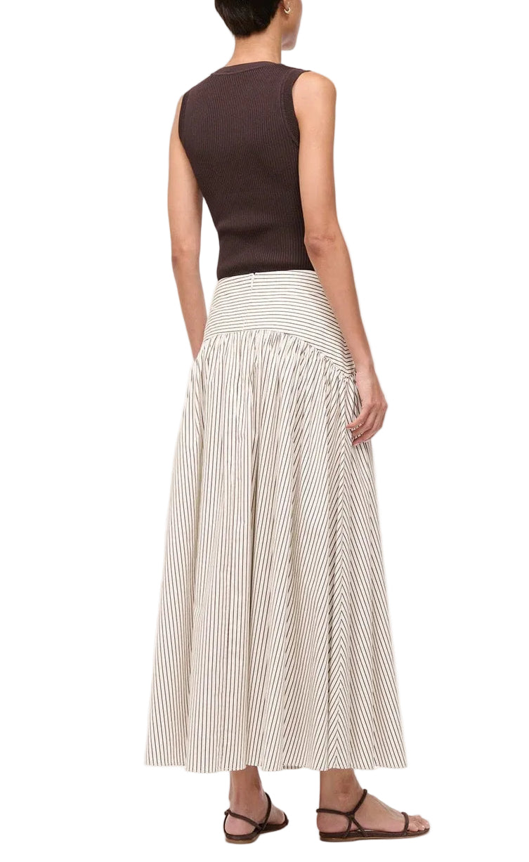 A person stands with their back to the camera, wearing a sleeveless dark brown top and the Staud Procida Skirt—a high-waisted white and beige striped maxi skirt with a curved yoke—paired with brown flat sandals.