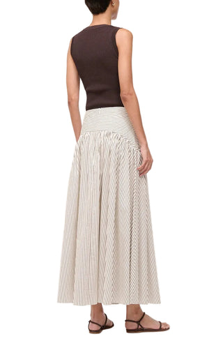 A person stands with their back to the camera, wearing a sleeveless dark brown top and the Staud Procida Skirt—a high-waisted white and beige striped maxi skirt with a curved yoke—paired with brown flat sandals.