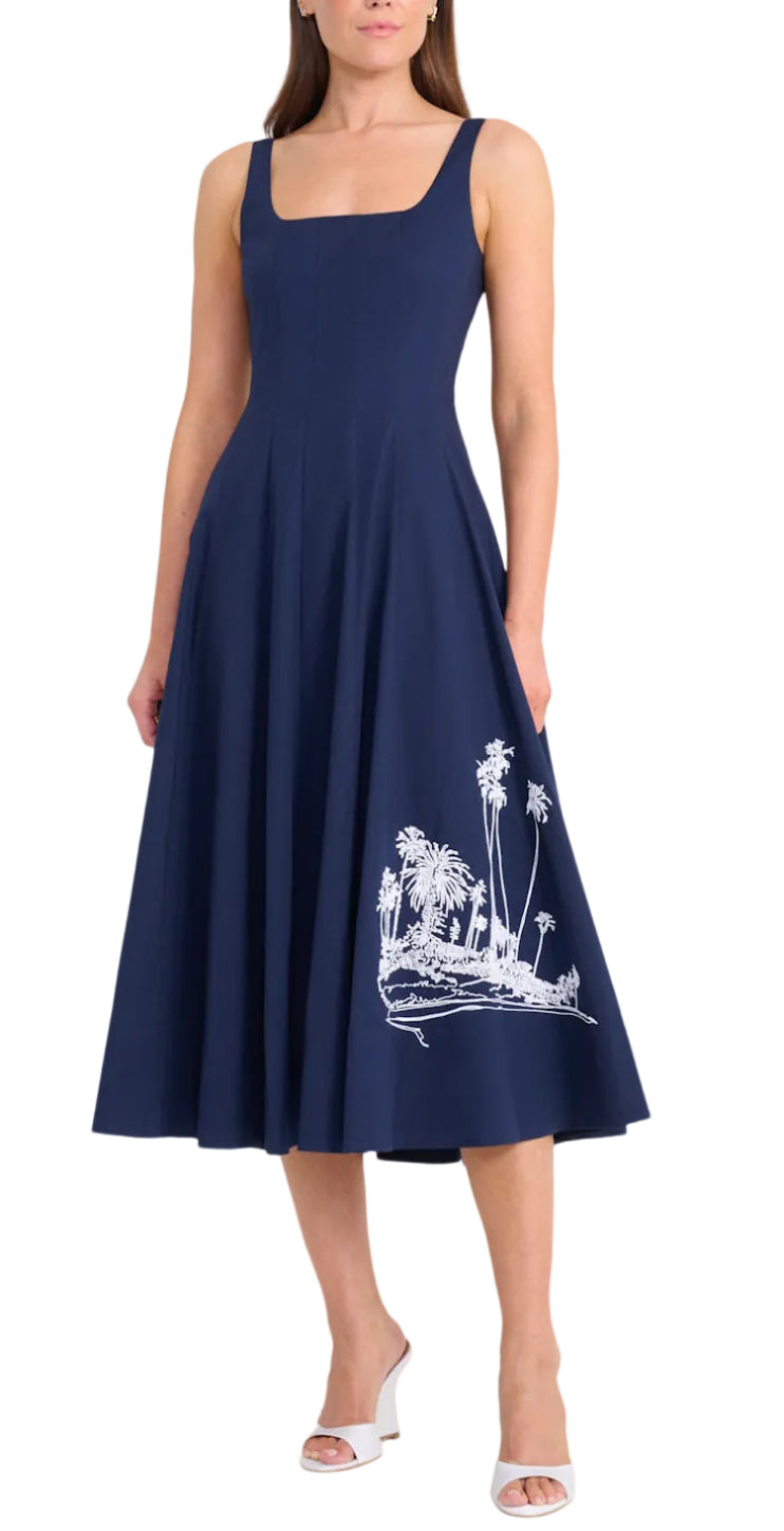 A woman wears the Staud Wells Island Embroidery Dress, a sleeveless navy cotton poplin piece with a square neckline and white palm tree embroidery near the hem, paired with white open-toe heels.