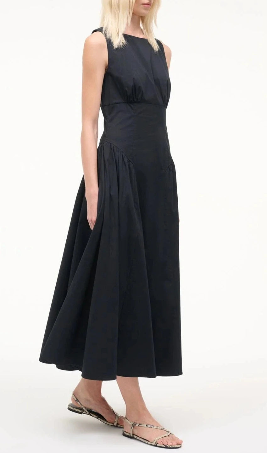 A woman wears the Staud Gwen Dress, a sleeveless, ankle-length A-line style with a fitted bodice and flared skirt, paired with flat snake-print sandals, standing against a plain white background.
