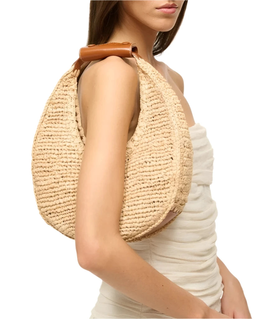 A person is wearing the Staud Moon Raffia Tote Bag, perfectly complementing their white, sleeveless, ruched dress.