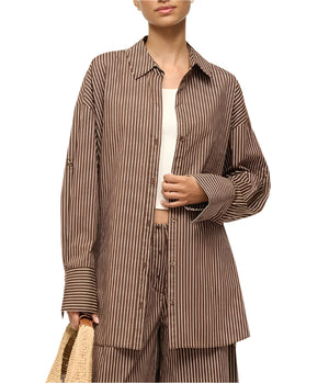 A person wearing the Staud Sandbar Top, a stylish two-piece set with brown and white stripes, carries a straw bag. The boxy button-up shirt is partially open, revealing a white top underneath.