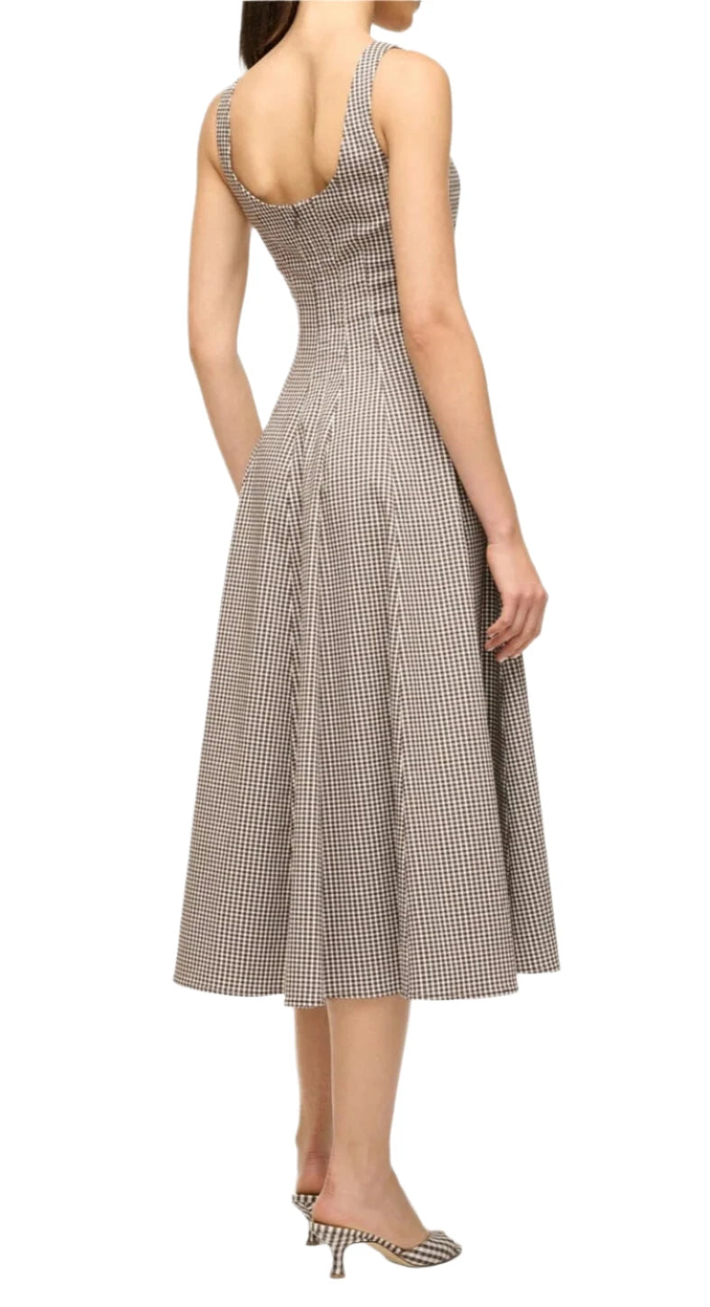 A woman wears the Staud Wells Micro Check Dress, a sleeveless, corset-style design with a fitted bodice and flared skirt. She pairs it with matching pointed heels, facing away.