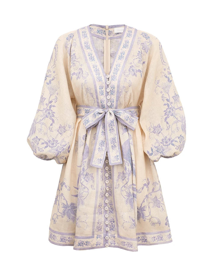 The Indra Plunge Mini Dress by Zimmermann features cream linen with lavender florals, long puffed sleeves, a plunging V-neck, front buttons, and a matching belted waist.