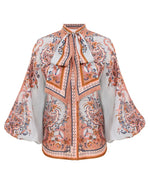 Zimmermann Aster Tie Neck Blouse featuring blue paisley floral patterns, wide sleeves, and a large neck bow, shown on a white background.