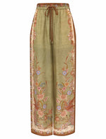 The Indra Relaxed Pant by Zimmermann features wide-leg silk habutae with a green base, brown drawstring waist, and floral patterns on sides and hem.