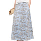 A woman in the Staud Axel Skirt, a blue floral a-line maxi, pairs it with a white top, woven clutch, and beige wedge sandals.