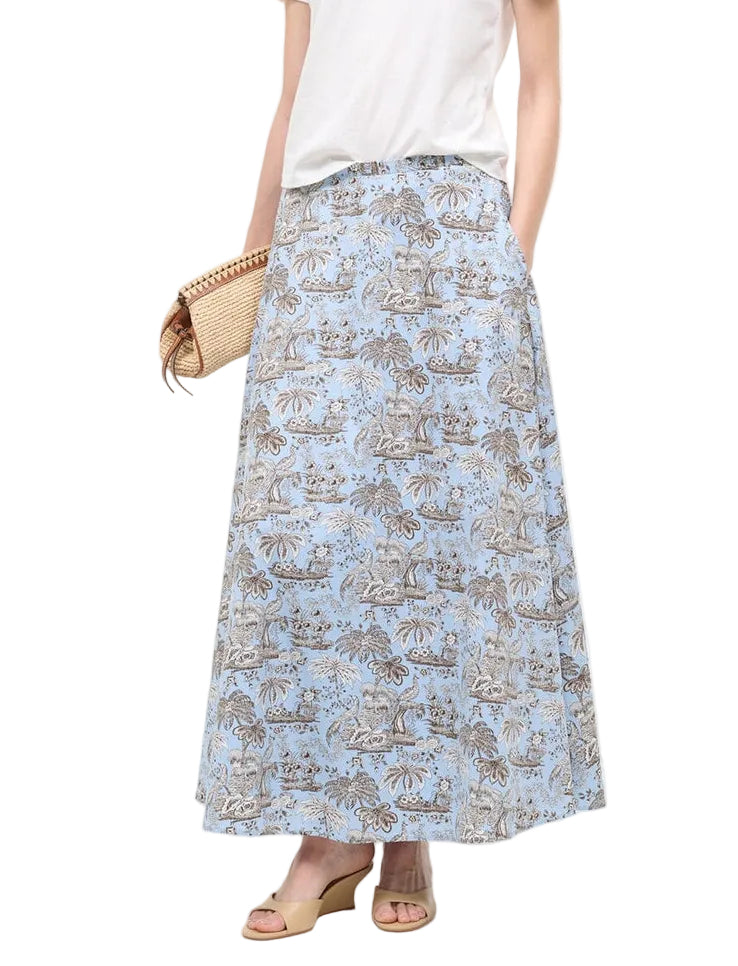 A woman in the Staud Axel Skirt, a blue floral a-line maxi, pairs it with a white top, woven clutch, and beige wedge sandals.