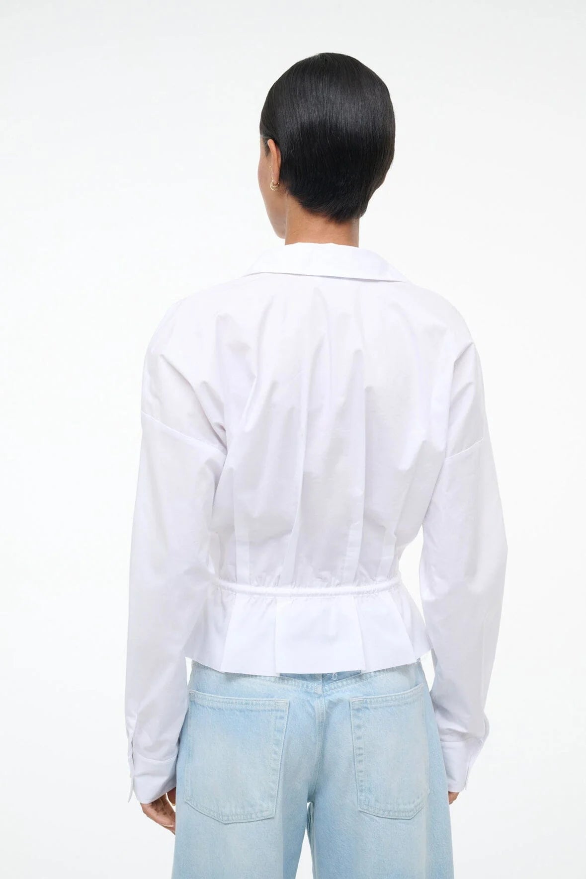 Person stands with their back to the camera, wearing the Staud Primrose Shirt—a white poplin button-down—paired with light blue jeans, set against a plain background.