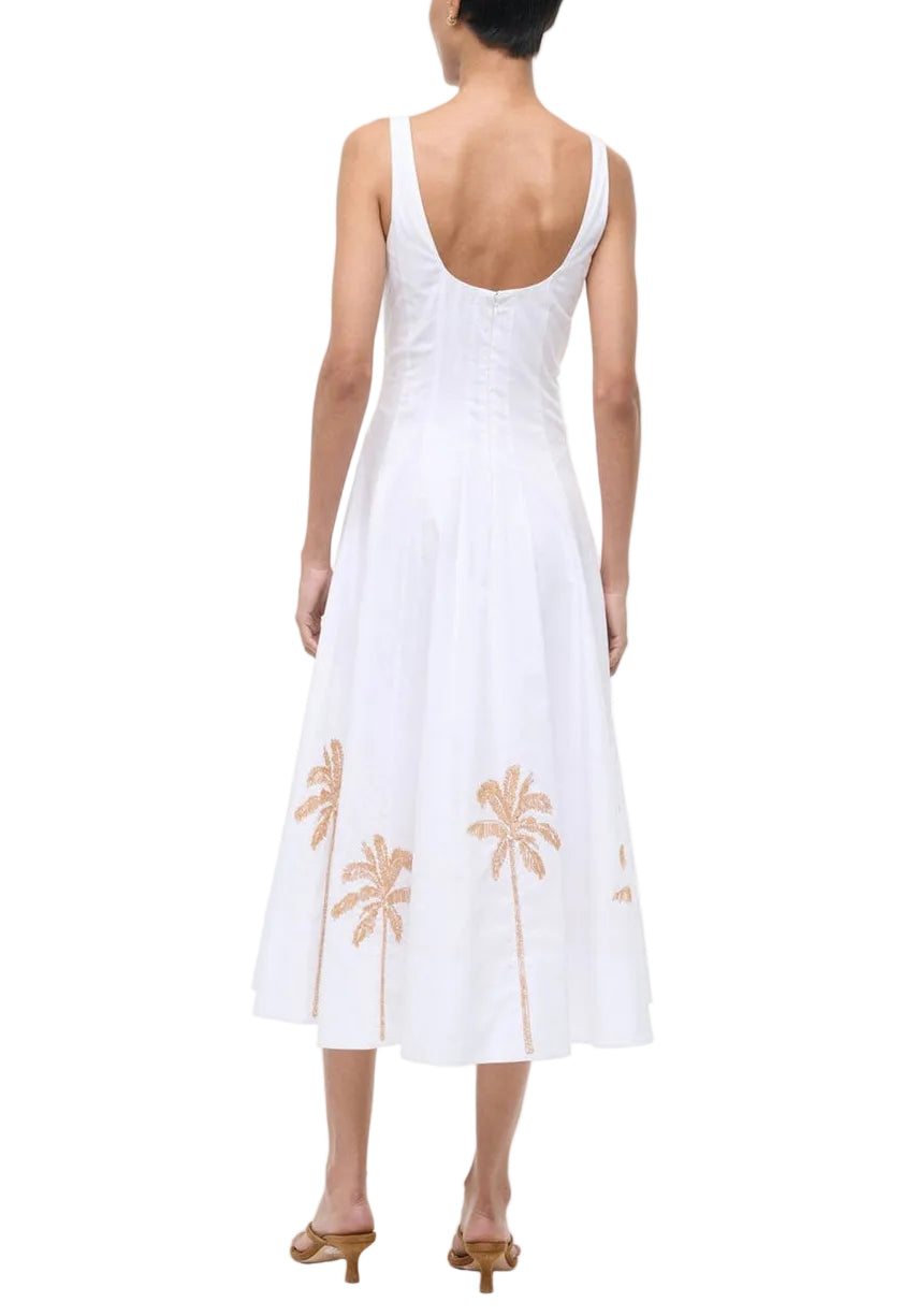 A person wearing the Staud Wells Palm Embroidery Dress—a sleeveless white cotton poplin dress with a fitted waist and tan palm tree embroidery near the hem—paired with heeled sandals.