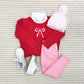 A cozy kids’ outfit featuring The Beaufort Bonnet Company’s Richmond Red Baby and Kids' Crawford Crewneck with a white bow, paired with light pink leggings, a pink pom-pom knit hat, and white shoes, laid out on textured carpet.