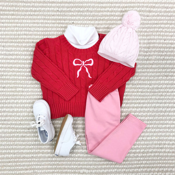 A cozy kids’ outfit featuring The Beaufort Bonnet Company’s Richmond Red Baby and Kids' Crawford Crewneck with a white bow, paired with light pink leggings, a pink pom-pom knit hat, and white shoes, laid out on textured carpet.