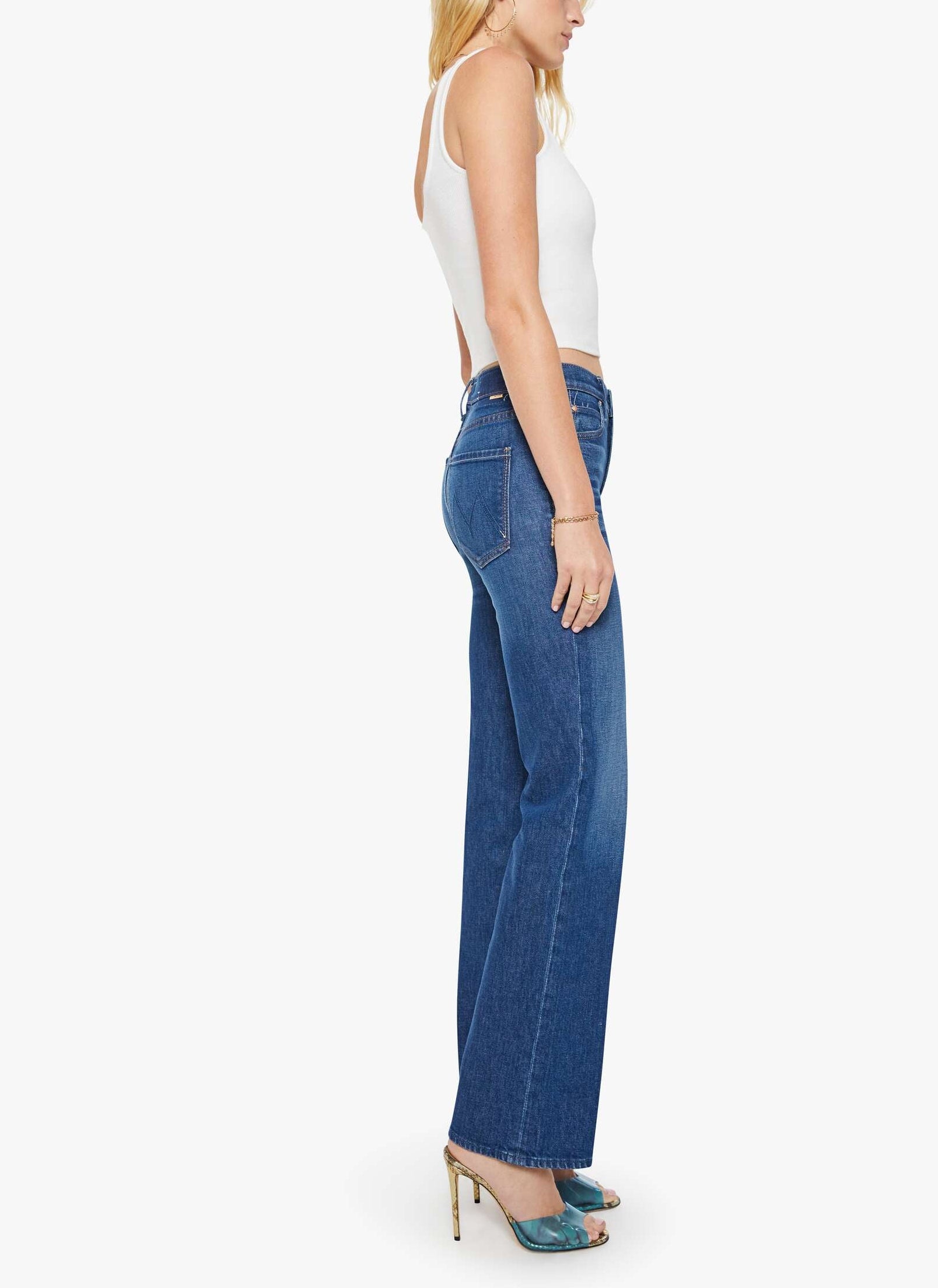 A woman stands sideways against a plain white background wearing the Mother Kick It high-rise straight-leg jean—crafted from recycled stretch denim with a 32-inch inseam—paired with a white sleeveless top and teal open-toe heels.