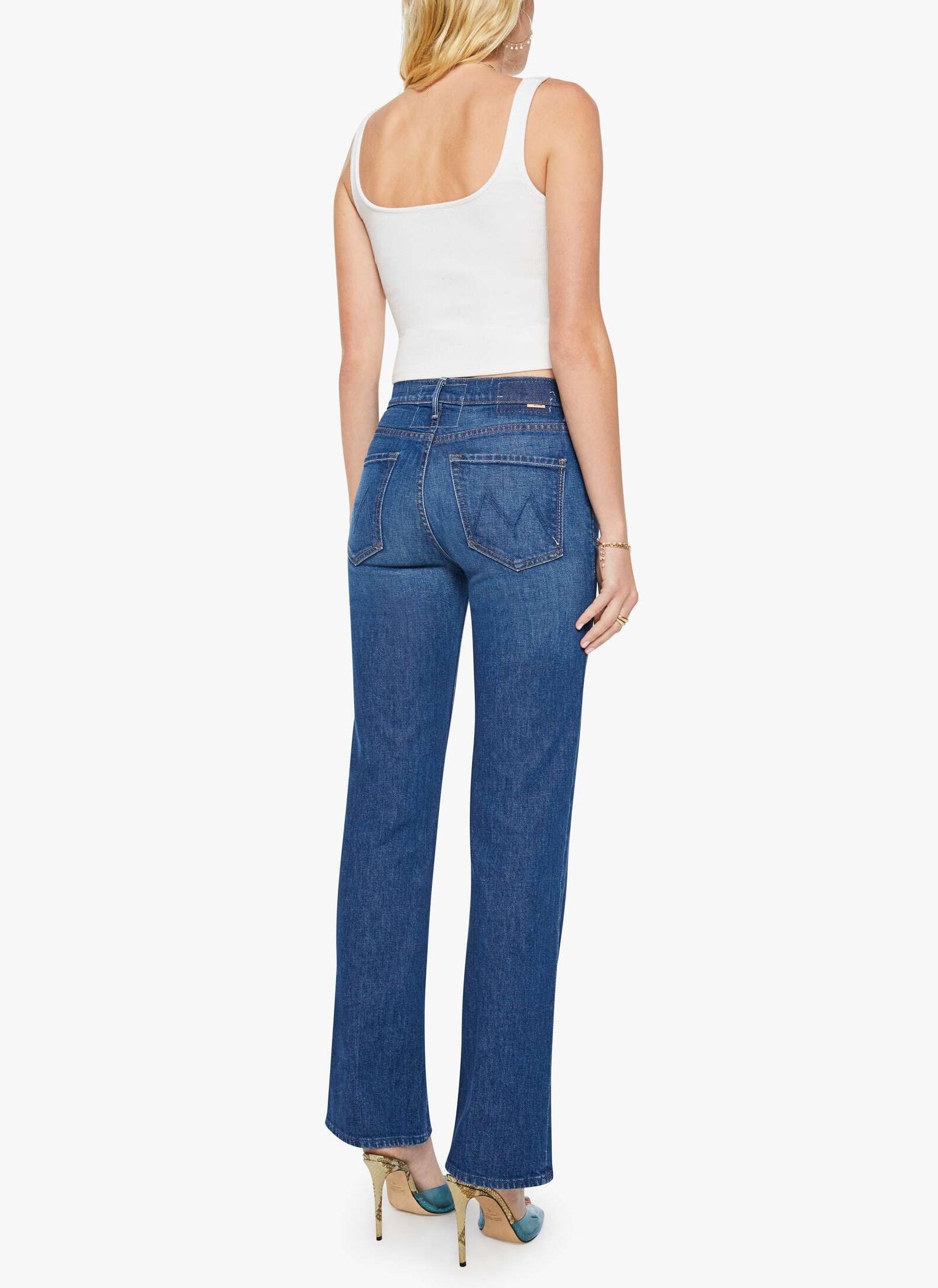 A woman stands facing away against a plain white background, wearing Mother’s Kick It blue high-rise straight-leg jeans crafted from recycled stretch denim with a 32-inch inseam, paired with a white tank top and beige heels.