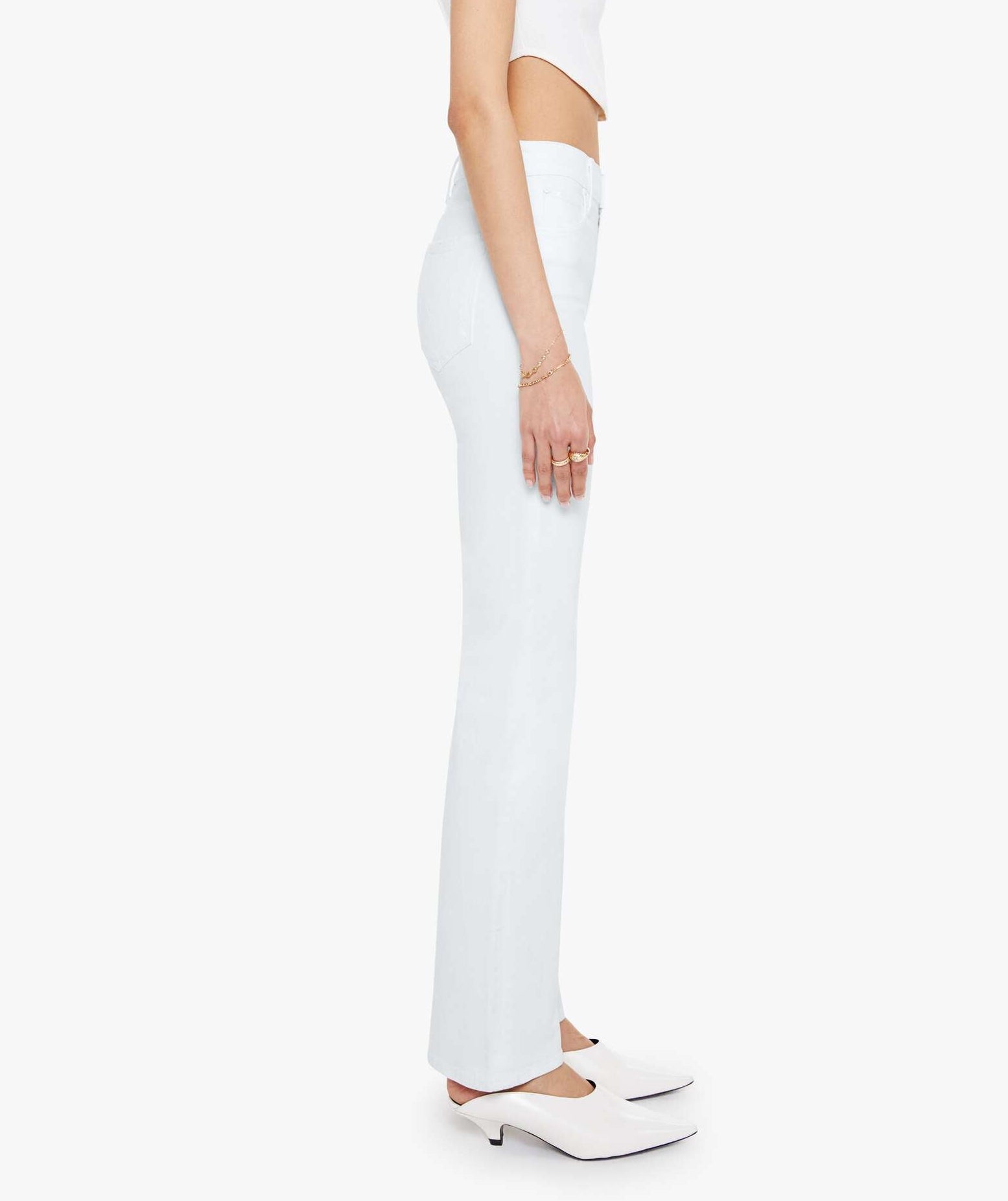 A person wearing the Mother Kick It high-rise straight-leg jeans, crafted from recycled stretch denim in a light mid-blue wash, pairs them with a white crop top and white heeled shoes while standing in profile against a plain white background.