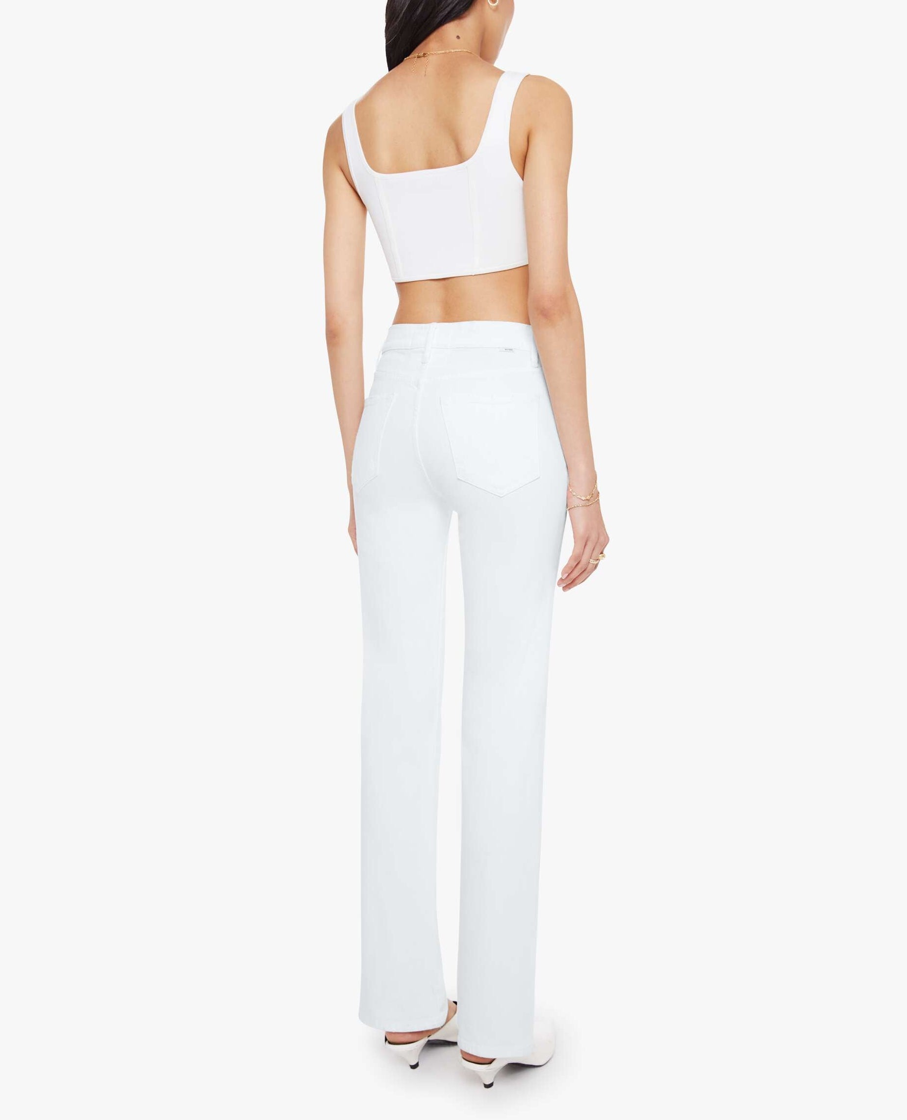 A woman stands facing away, wearing Mother’s Kick It high-rise straight-leg jeans crafted from recycled stretch denim, a white crop top, and white heels against a plain white background.