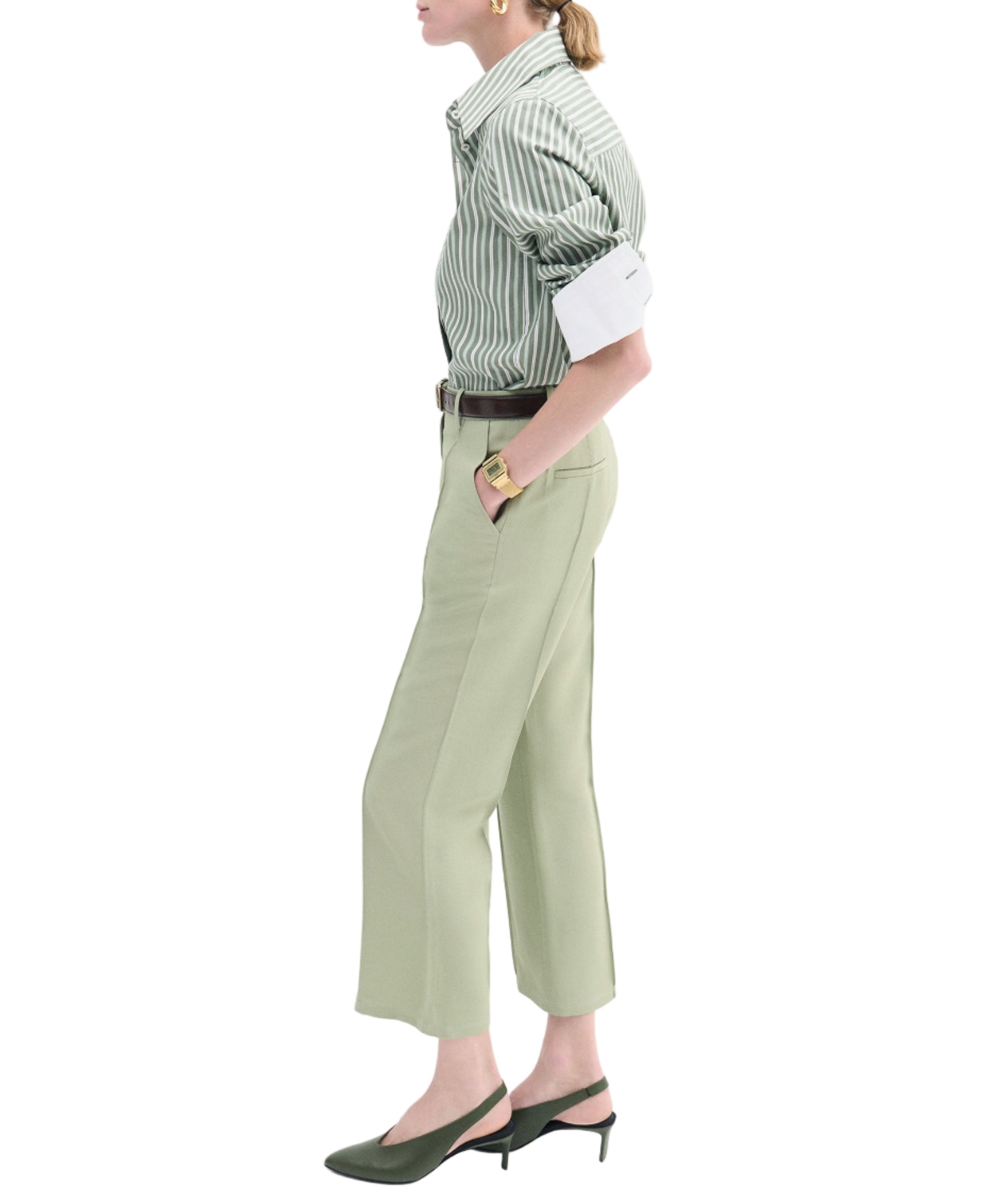 A person in a CO Fitted Classic Shirt in green cotton poplin with stripes, paired with light green tailored cropped pants, green slingback heels, and gold jewelry, stands in side profile.