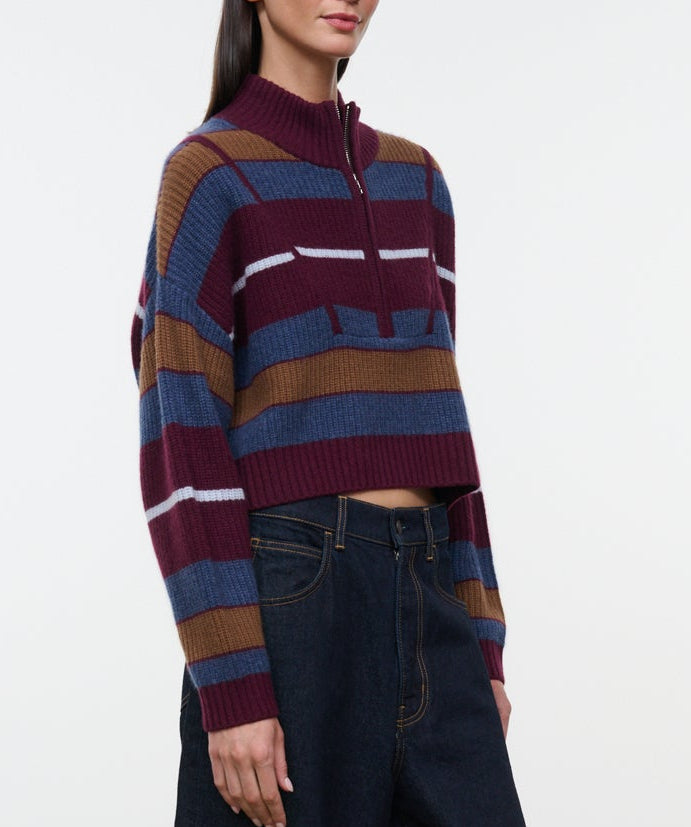 A woman wearing the Staud Hampton Cashmere Sweater—a cropped, long-sleeve, striped style with a half-zip collar—paired with high-waisted dark jeans, stands against a plain white background.