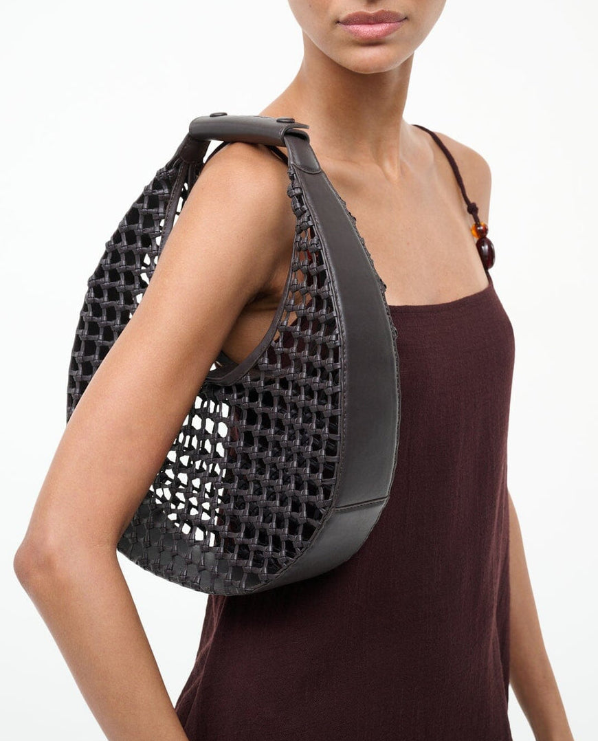 A woman in a sleeveless brown dress carries the Staud Moon Hand-Knotted Tote Bag, a large black leather crescent-shaped tote with solid leather edges, on her shoulder.