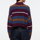 A person is shown from the back wearing the Hampton Cashmere Sweater by Staud—a ribbed, long-sleeve sweater featuring burgundy, blue, brown, and white horizontal stripes in a chunky cashmere knit—paired with dark denim jeans.