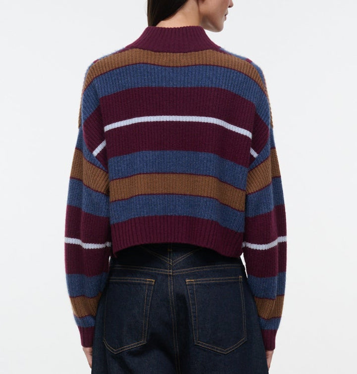 A person is shown from the back wearing the Hampton Cashmere Sweater by Staud—a ribbed, long-sleeve sweater featuring burgundy, blue, brown, and white horizontal stripes in a chunky cashmere knit—paired with dark denim jeans.