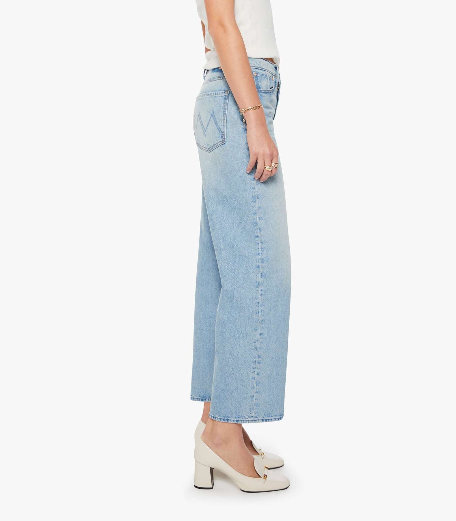 A person stands sideways against a plain background wearing The Dodger Flood wide-leg cropped jeans by Mother in light blue denim, paired with a white short-sleeve top and white heeled shoes.