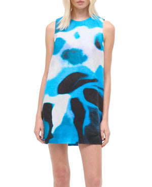 A woman stands against a white background wearing the Staud Brea Linen Dress, a sleeveless mini with a blue, black, and white abstract print, styled with a breathable layering piece for comfort.