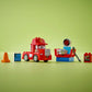 The LEGO® DUPLO® Mack at the Race set by Legos - Toyhouse features a red truck, orange cone, blue screen, engine, tire, wrench, and decal toolbox arranged on a green surface with a green background.