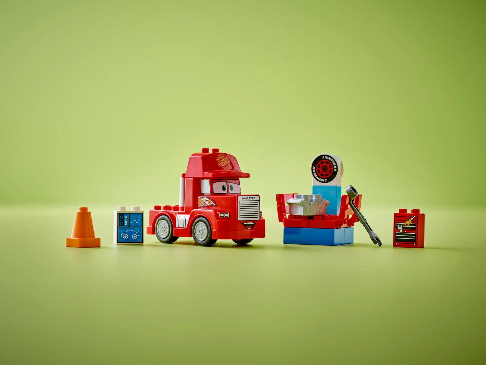 The LEGO® DUPLO® Mack at the Race set by Legos - Toyhouse features a red truck, orange cone, blue screen, engine, tire, wrench, and decal toolbox arranged on a green surface with a green background.