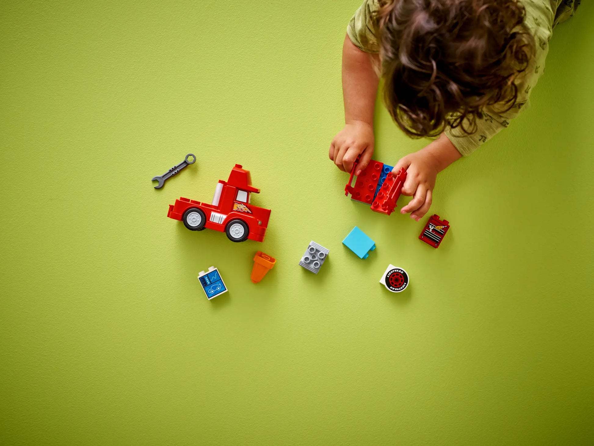 A child assembles LEGO® DUPLO® Mack at the Race blocks from Legos - Toyhouse on a green surface, with pieces and a toy wrench scattered nearby.