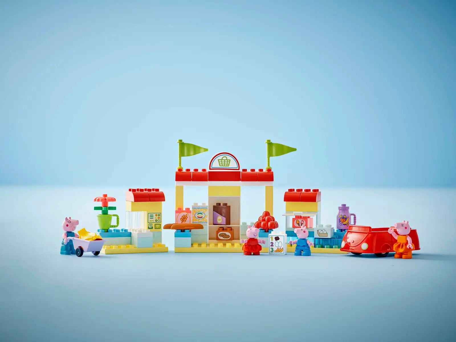 The LEGO® DUPLO® Peppa Pig Supermarket by Legos - Toyhouse features a building with flags, toy food items, small figures, a cart, and a red car—perfect for preschool kids.
