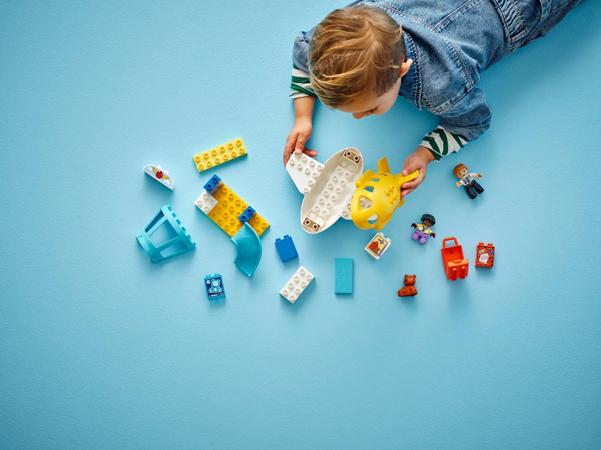 A young child in denim plays on a blue surface with colorful preschool toys, including building blocks, animal and vehicle figures, and the LEGO® DUPLO® First Time at the Airport set by Legos - Toyhouse.