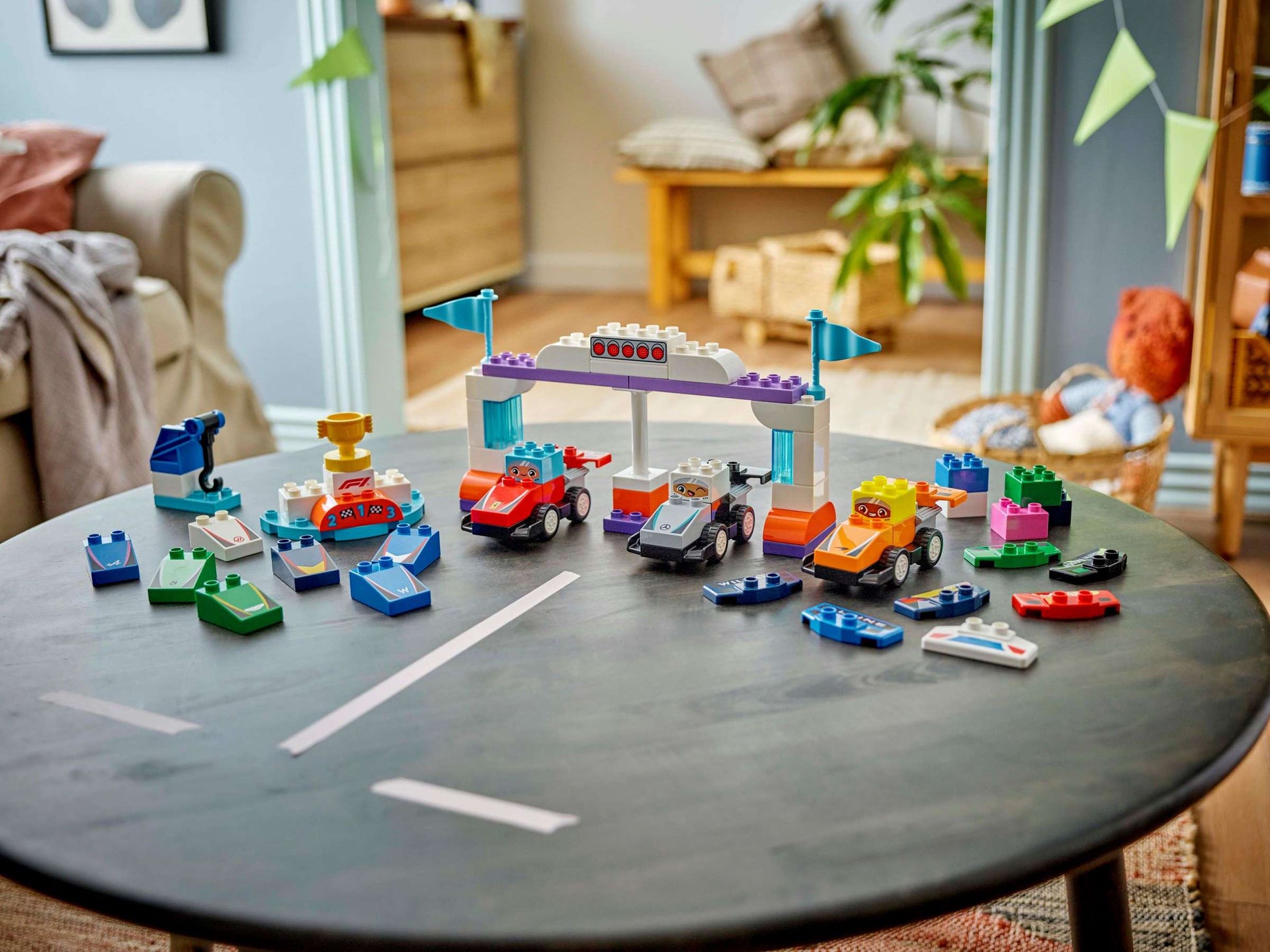 A round black table displays Legos - Toyhouse LEGO® DUPLO® F1® Team Race Cars & Drivers, with a living room and green plants in the background.