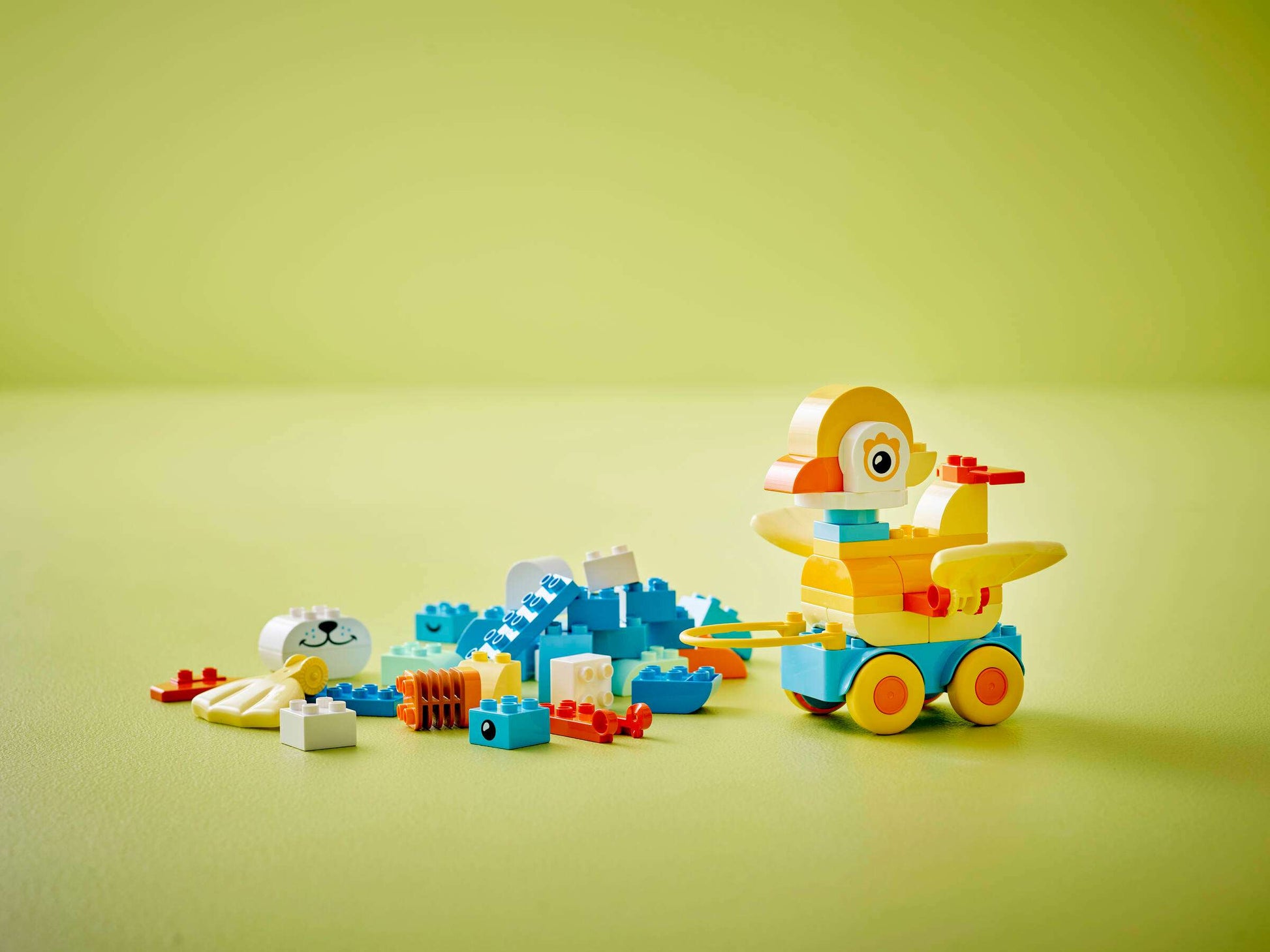 The LEGO® DUPLO® 3in1 Animals on Wheels by Legos - Toyhouse includes colorful blocks and animal figures with wheels—such as a yellow duck—plus blue, white, and orange pieces, all displayed on a light green surface with a green background.