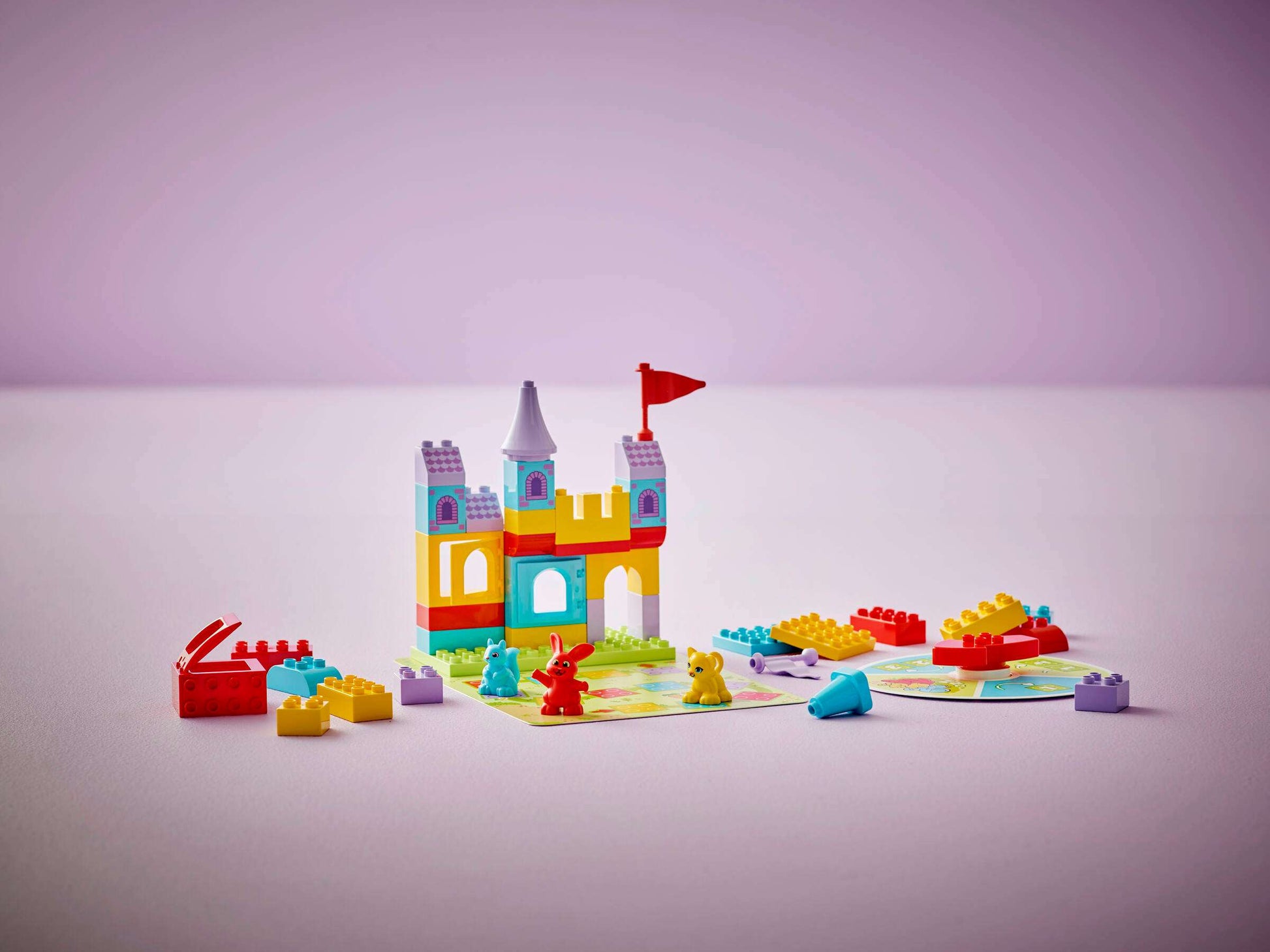 The LEGO® DUPLO® Hopsy's Castle Game by Legos - Toyhouse features a colorful toy castle with a red flag, plastic animal figures, building blocks, and play cards on a pale purple surface for fun educational play.