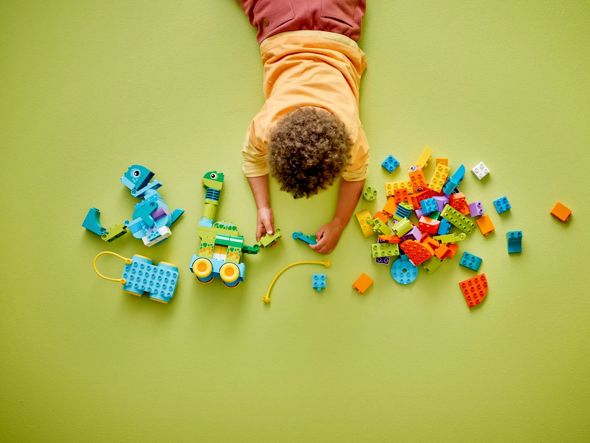 A child plays on a green floor with the LEGO® DUPLO® 3in1 Dinosaurs on Wheels set by Legos - Toyhouse, assembling colorful dinosaur pieces from this toddler building set.