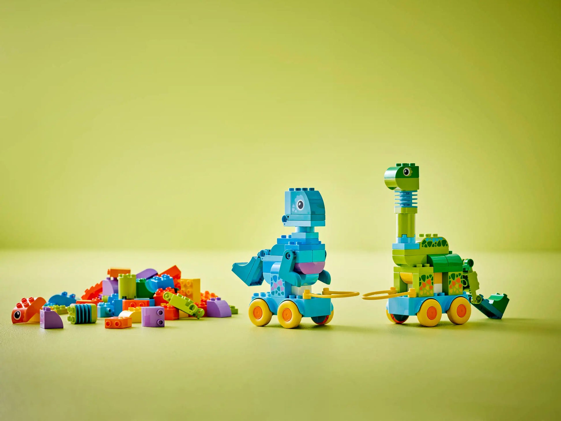 The LEGO® DUPLO® 3in1 Dinosaurs on Wheels by Legos - Toyhouse features two colorful dinosaur toys with wheels and loose bricks on the left, all set against a light green background.