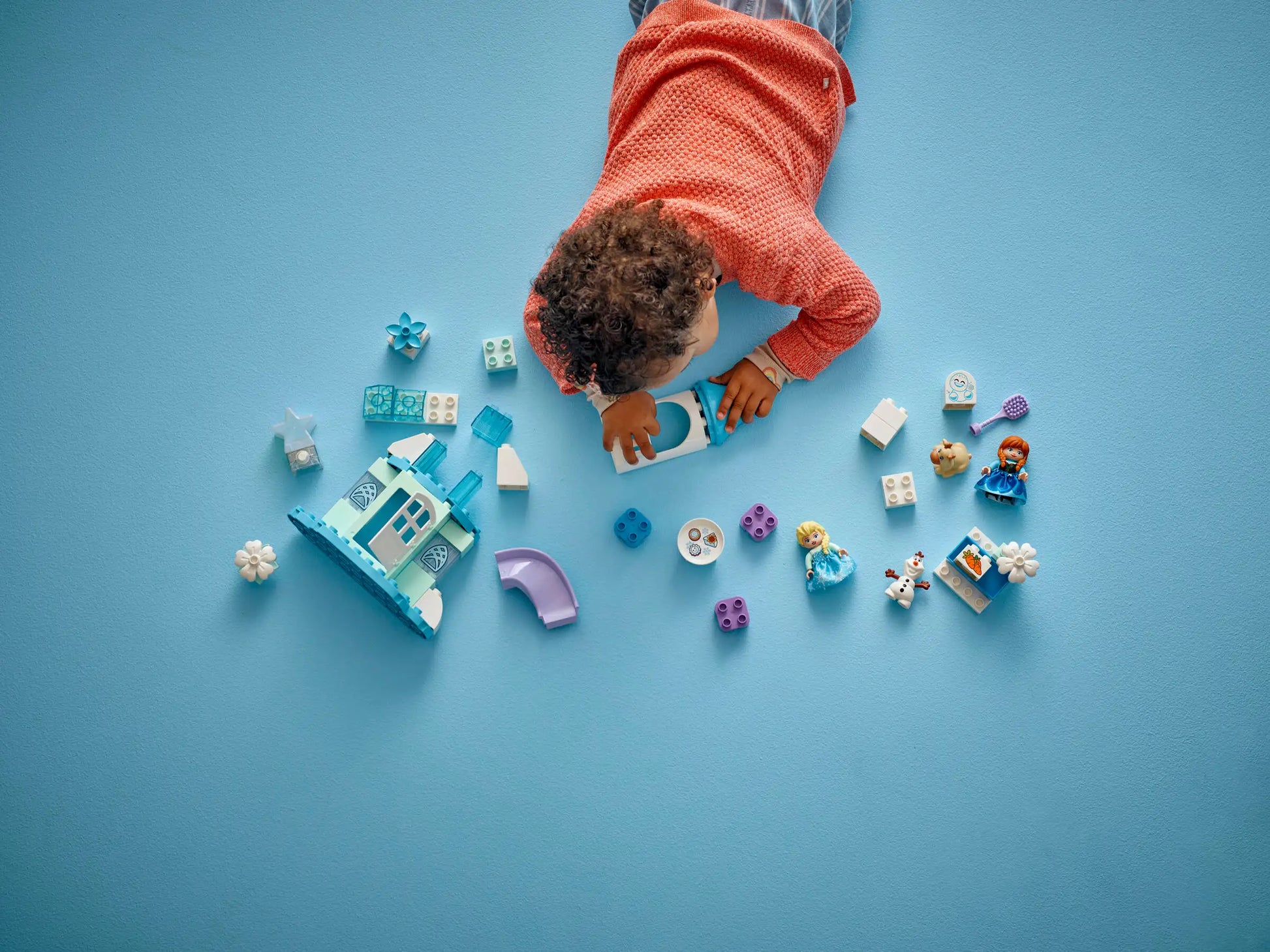 A child in a red sweater plays on a blue floor with Legos - Toyhouse pieces, including character figures and a toy house from the LEGO® DUPLO® Anna and Elsa's Frozen Castle Party set.