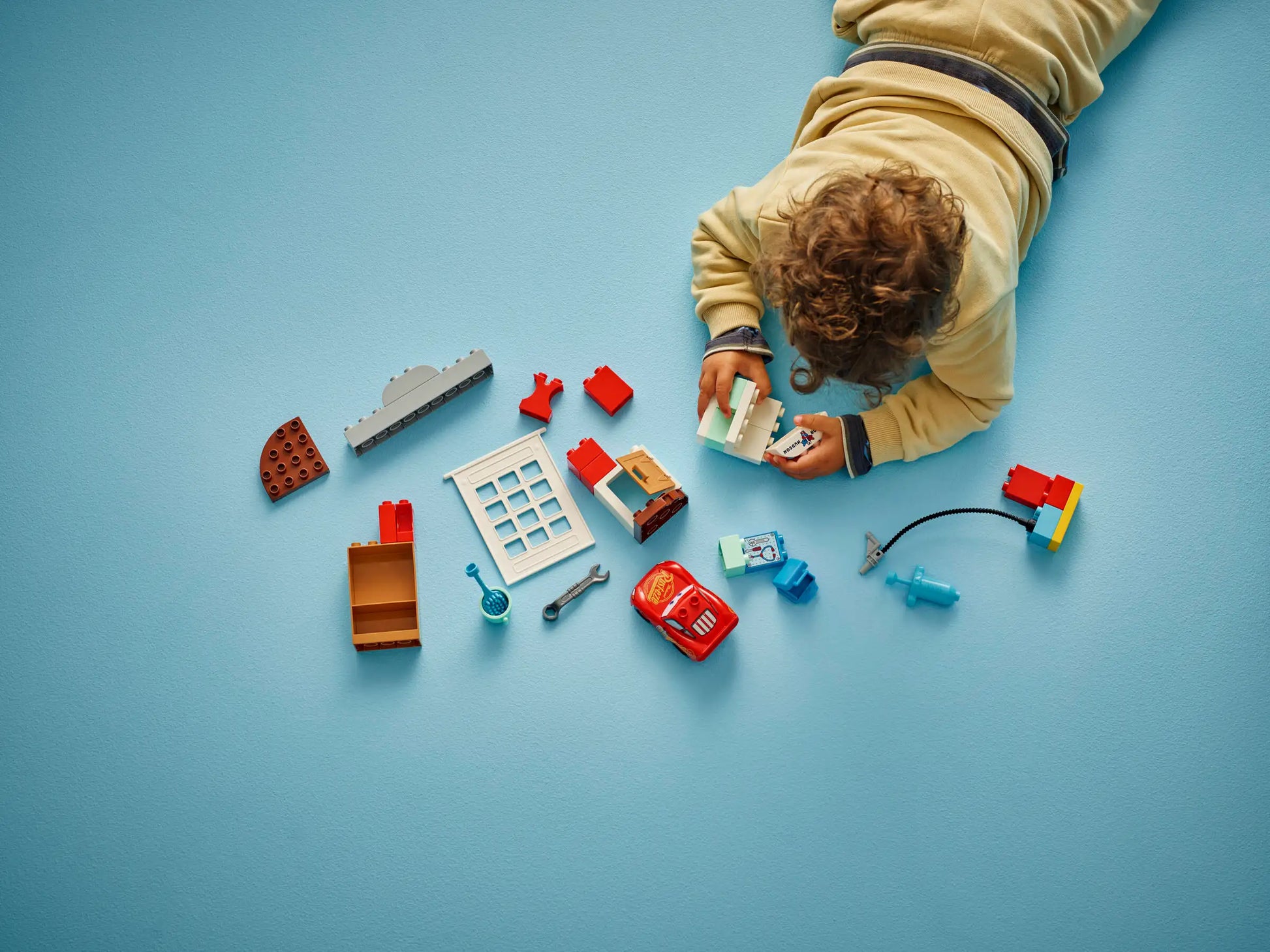 A child in beige clothes lies on a light blue floor, playing with the LEGO® DUPLO® McQueen's Visit to Doc's Garage set from Legos - Toyhouse.