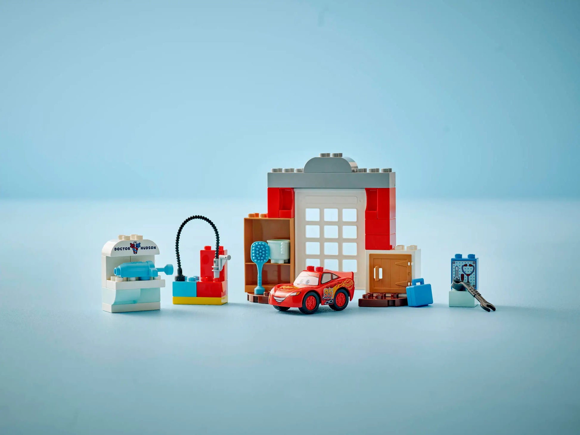 The LEGO® DUPLO® McQueen's Visit to Doc's Garage by Legos - Toyhouse features a buildable race car toy displayed with garage accessories and buildings on a light blue background.