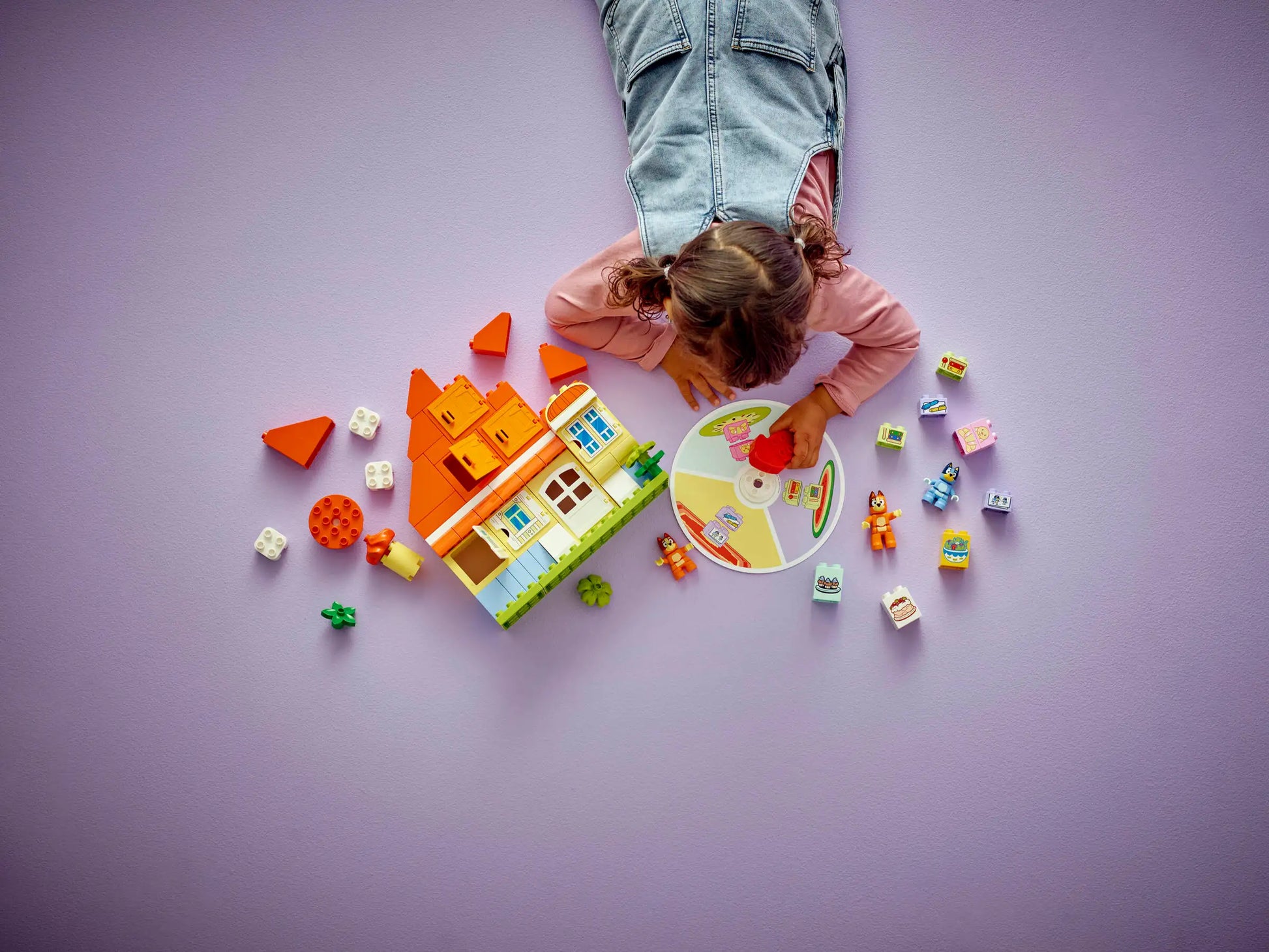 A child plays on a purple floor with the LEGO® DUPLO® Bluey's Family House with Memory Game by Legos - Toyhouse, featuring building blocks, a colorful spinning memory game board, and small toy figures and pieces.