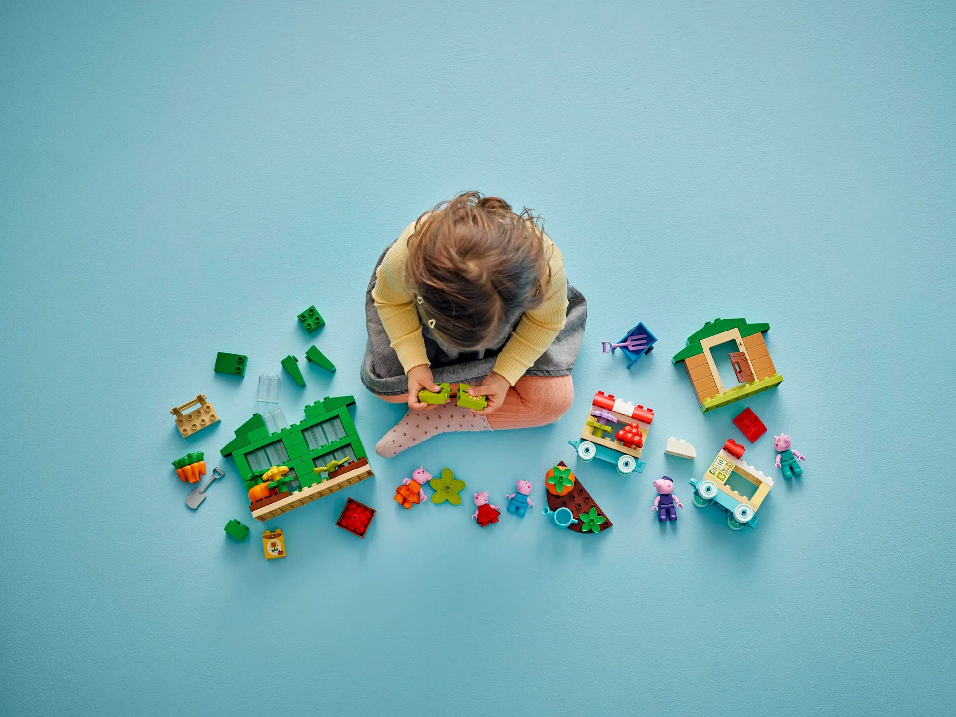 A child sits on a blue floor, playing with the LEGO® DUPLO® Grandpa Pig's Garden and Greenhouse building set from Legos - Toyhouse, arranging colorful blocks and Peppa Pig figures in front of them.