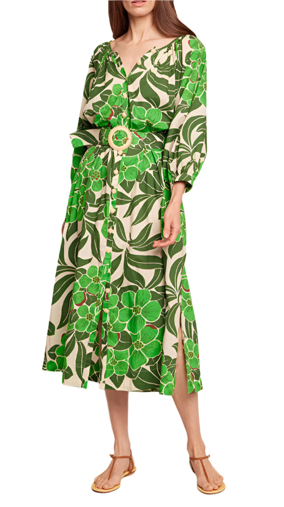 The woman wears an elegant Cara Cara Simone Dress, showcasing a green and beige floral print with a belted waist and crochet-covered buttons, perfectly paired with sandals.