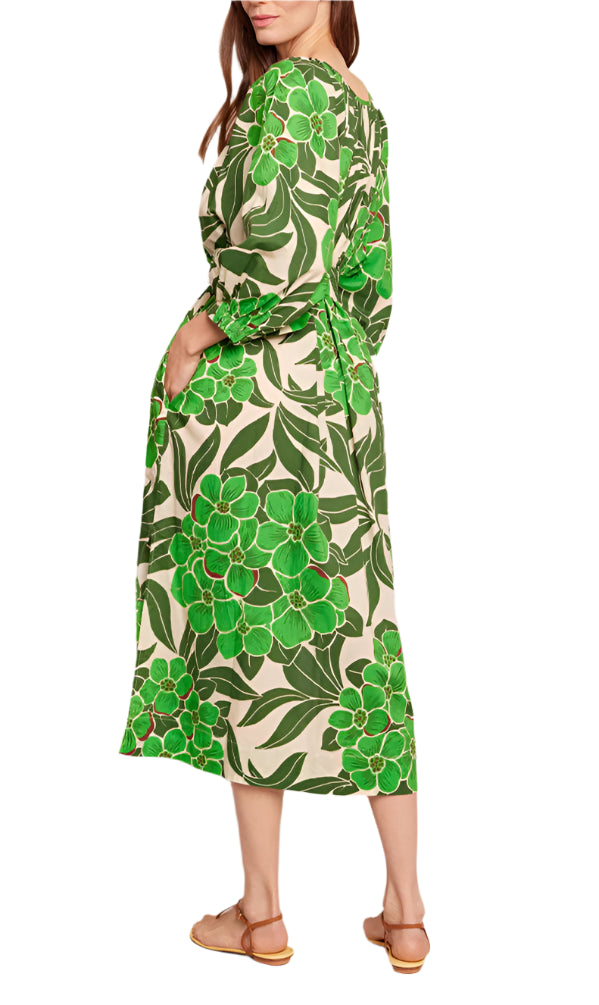 The woman stands sideways in a green floral Cara Cara Simone Dress, highlighting crochet-covered buttons, and pairs it with tan sandals.