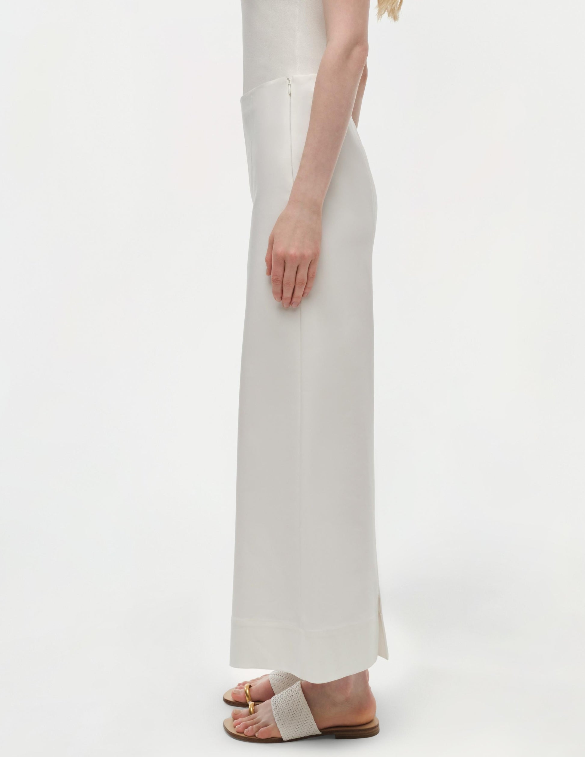 A person stands in profile against a plain white background, wearing the Simkhai Kallin Cropped Wide Leg Pant in white cotton, a sleeveless white top, and white sandals with gold buckle detail.