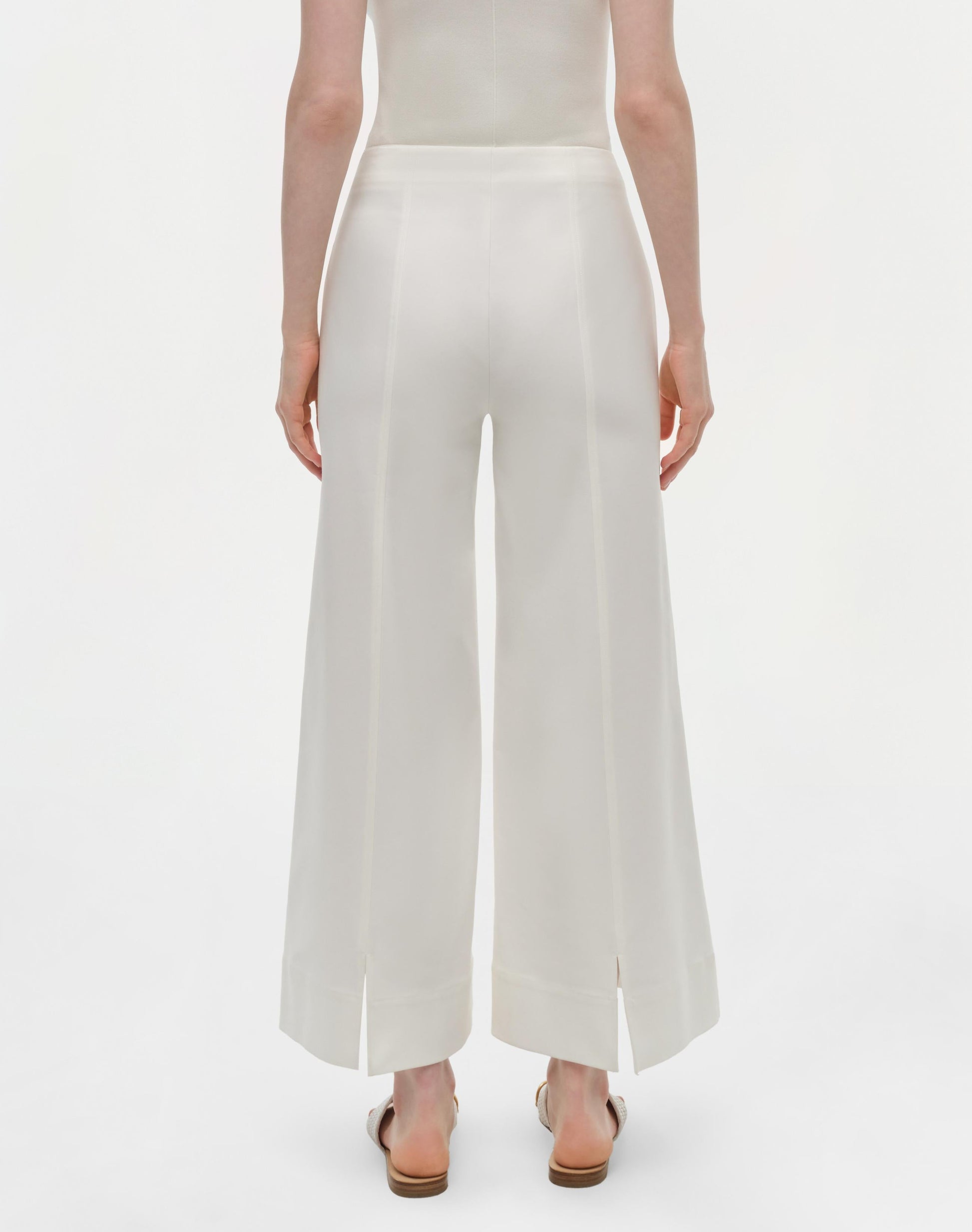 A person faces away, wearing Simkhai's Kallin Cropped Wide Leg Pant in white—high-waisted, heavy cotton trousers with front hem slits—paired with sandals, against a plain white background.