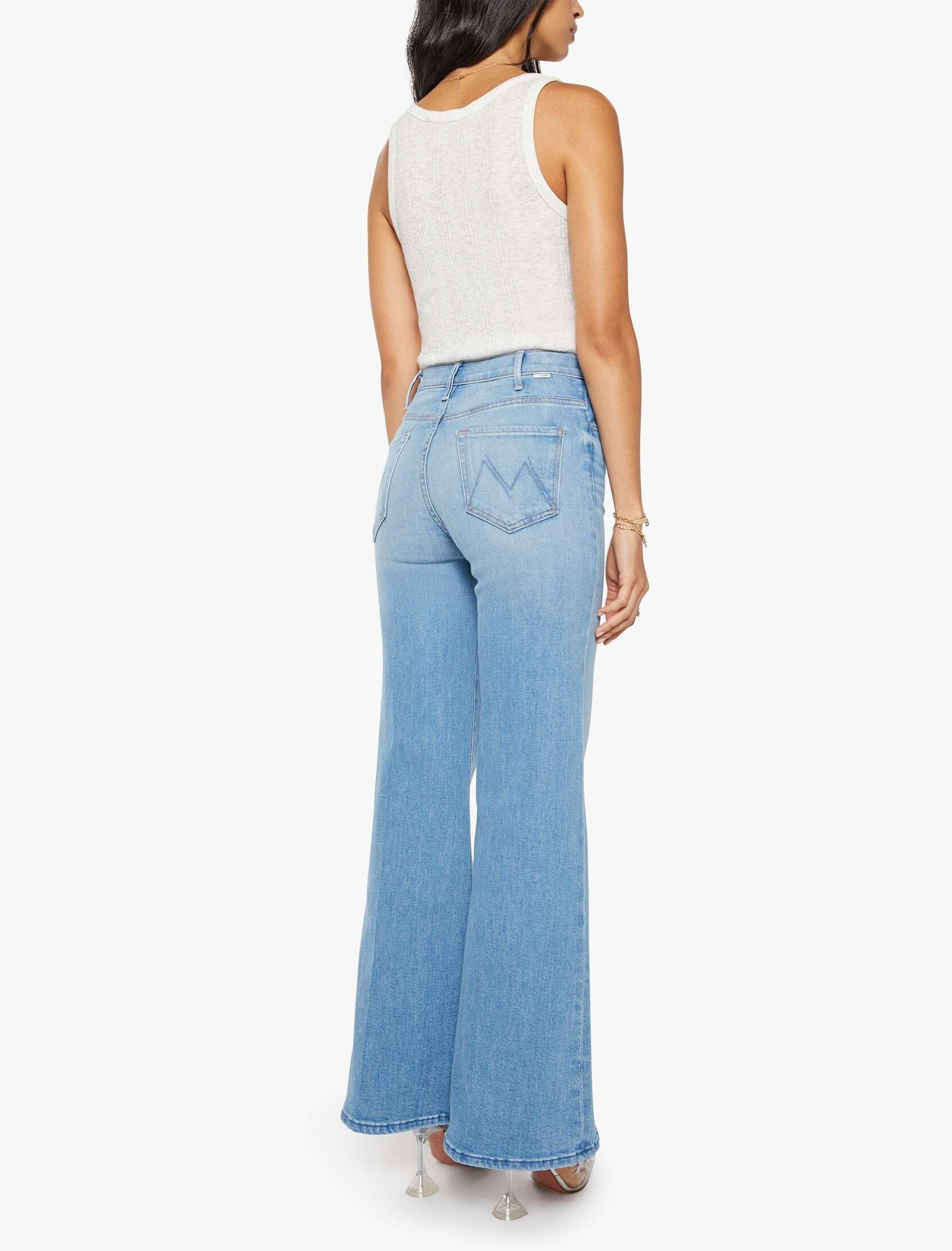 A woman is standing with her back to the camera against a white background, wearing a white sleeveless top and The Hustler Roller Sneak high-waisted stretch denim wide leg jeans by Mother in light blue.